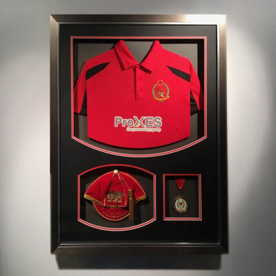 A red sports shirt, cap and medal in three separate apertures, on black and red mount in a pewter frame.