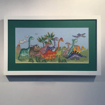 A colourful dinosaur cross stitch on a green mount in a white frame.