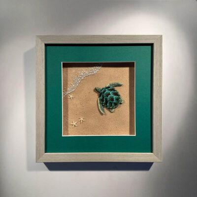 a turtle made from textiles in a window surrounded by green mount board in a pale wooden frame
